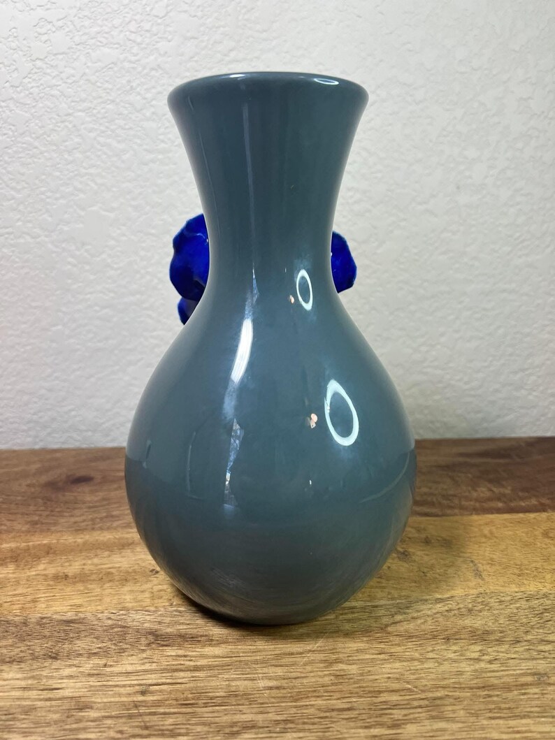 May include: A gray-blue ceramic vase with a rounded body and flared neck. A vibrant blue flower is visible behind the vase. The vase is displayed on a wooden surface.