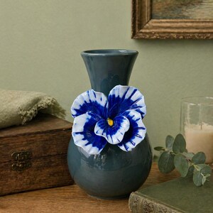 May include: A teal ceramic vase with a large, decorative blue and white flower attached. The flower has a yellow center. The vase is on a wooden surface, with a wooden box and a green book nearby. A painting hangs in the background.