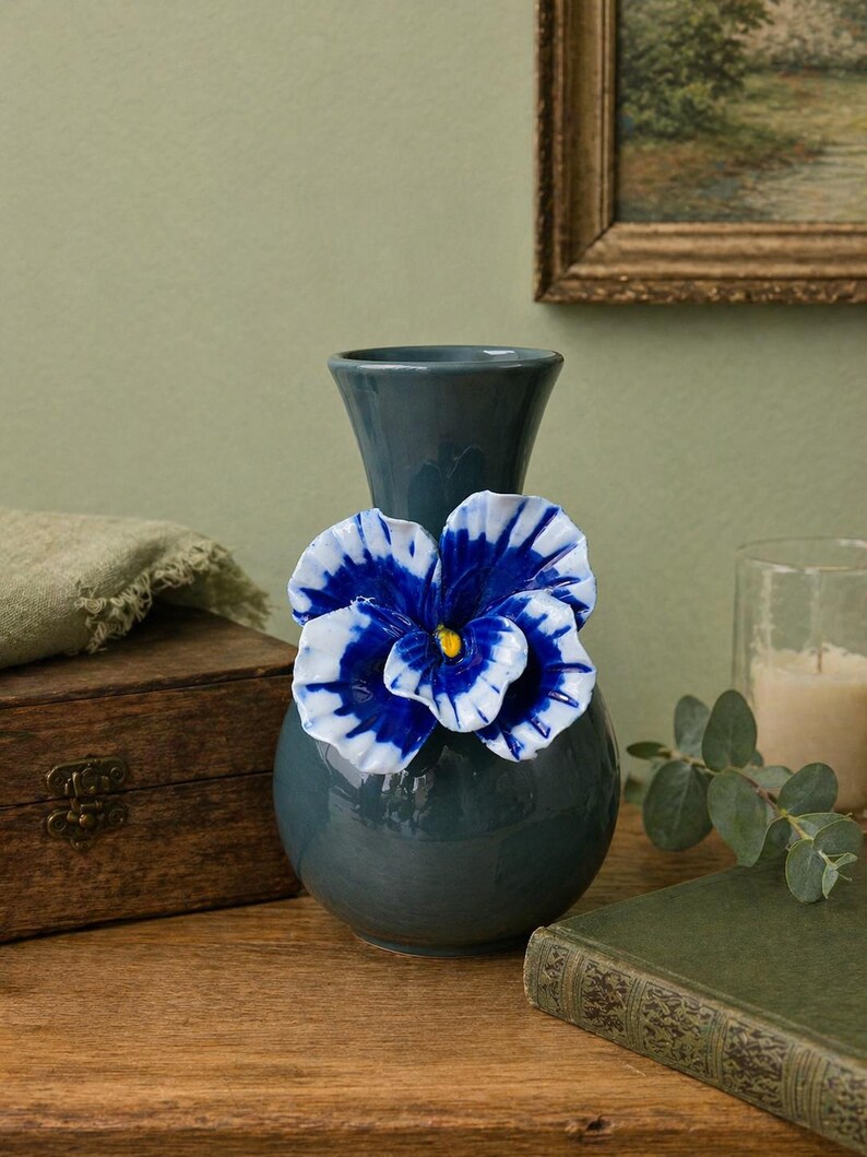 May include: A teal ceramic vase with a large, decorative blue and white flower attached. The flower has a yellow center. The vase is on a wooden surface, with a wooden box and a green book nearby. A painting hangs in the background.