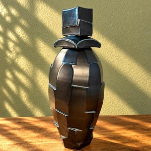 May include: A tall, dark charcoal-coloured ceramic vessel with a faceted, geometric surface texture. The object features a matching lid with a square top and a curved base, creating a segmented, layered appearance across the entire body.