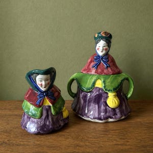 May include: A pair of whimsical ceramic figurines, one a small creamer and the other a sugar bowl. Both are painted with a lady design, featuring a red cape, green shawl, and purple skirt. The sugar bowl has handles and a lid.