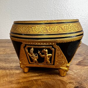 May include: A black and gold decorative bowl with ornate detailing. The bowl features gold trim and accents, with a detailed relief of figures on the front. The bowl is set on a wooden surface.