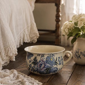 May include: A ceramic chamber pot featuring a cream-colored base with a detailed blue and purple floral pattern. The vessel has a wide, circular opening, a curved handle on the right side, and a rounded body decorated with intricate botanical motifs.