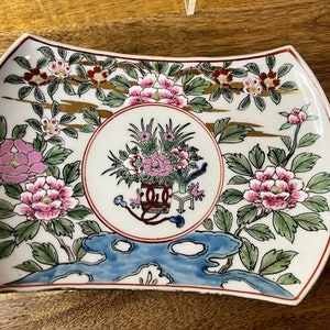 May include: A white ceramic plate with a scalloped edge features a floral design in pink, green, and blue. The center of the plate has a circular design with a floral arrangement in a basket.