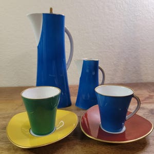 May include: A set of colorful ceramic coffee cups and saucers. The cups are blue, green, and red, and the saucers are yellow, red, and brown. The set also includes a blue and white ceramic creamer and a blue and white ceramic coffee pot.
