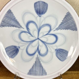 May include: White ceramic plate with a blue floral design. The plate features a central flower motif and stylized blue shapes. A thin pink line borders the edge of the plate.