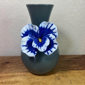 May include: A gray ceramic vase with a blue and white flower accent. The flower has a yellow center. The vase is on a wooden surface.