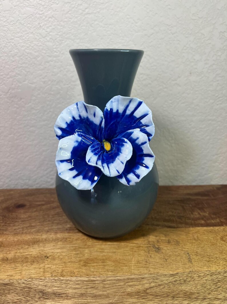 May include: A gray ceramic vase with a blue and white flower accent. The flower has a yellow center. The vase is on a wooden surface.
