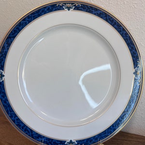 Puede incluir: Un plato de cena de porcelana blanca con un borde azul y dorado. El borde presenta un diseño azul marmoleado con detalles dorados y un motivo floral.