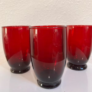 May include: Three red glass tumblers with a rounded base. The glasses are clear and have a slight reddish tint.