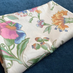 May include: Folded cream-colored fabric with a floral pattern. The design features large flowers in shades of pink, blue, orange, and green, with gold accents. The fabric is resting on a dark blue surface.