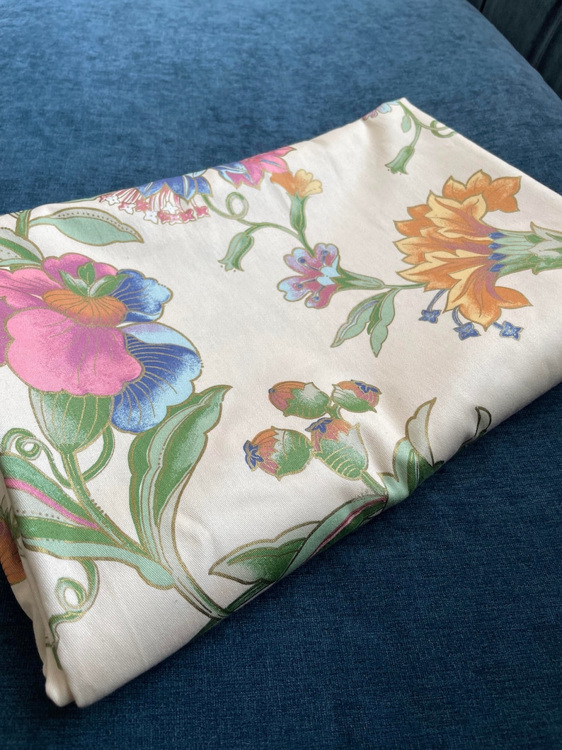 May include: Folded cream-colored fabric with a floral pattern. The design features large flowers in shades of pink, blue, orange, and green, with gold accents. The fabric is resting on a dark blue surface.