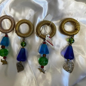 Puede incluir: Cuatro anillos de servilleta dorados con colgantes. Cada anillo tiene un colgante diferente: un elefante verde, un corazón, un pájaro verde y un corazón. Los colgantes son de metal y tienen un aspecto vintage.