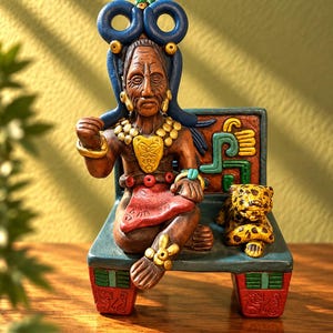 May include: A hand-painted clay figurine of a seated figure wearing a large blue headdress, gold-coloured jewellery, and a red loincloth. The figure sits on a rectangular bench next to a small yellow and black spotted leopard, with carved details.
