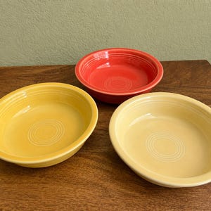 May include: Three round ceramic bowls featuring a concentric ring design on the interior base and rim. The set includes one bright orange bowl, one vibrant yellow bowl, and one pale yellow bowl, all with a smooth, glossy finish and curved edges.