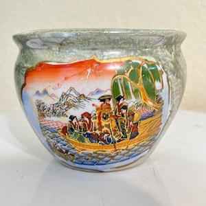 Könnte beinhalten: Ein grüner Keramiktopf mit einer japanischen Szene, die auf der Seite bemalt ist. Die Szene zeigt Menschen in einem Boot auf einem Fluss mit Bergen und Bäumen im Hintergrund.