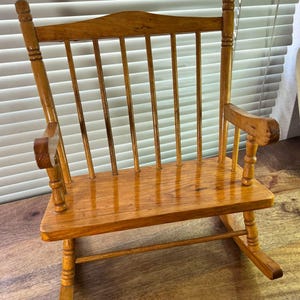 May include: A small, wooden rocking chair with a glossy finish. The chair has a slatted back, curved armrests, and a seat. The chair is made of light brown wood and is designed for a doll or small child.
