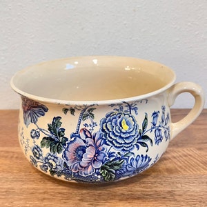 Puede incluir: Un orinal de cerámica blanca con un diseño floral azul y verde. El orinal tiene una asa en el lateral y un fondo redondeado.