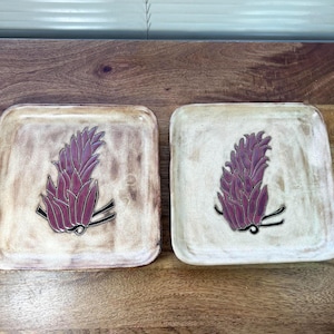 May include: Two square ceramic plates with a botanical design. Each plate features a maroon-colored floral motif with black accents. The plates have a cream and brown mottled background and are displayed on a wooden surface.