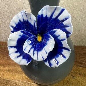 May include: A ceramic vase in a dark gray hue, adorned with a large, vibrant blue and white flower. The flower's petals display a striking contrast of colors, with a yellow center. The vase sits on a wooden surface.