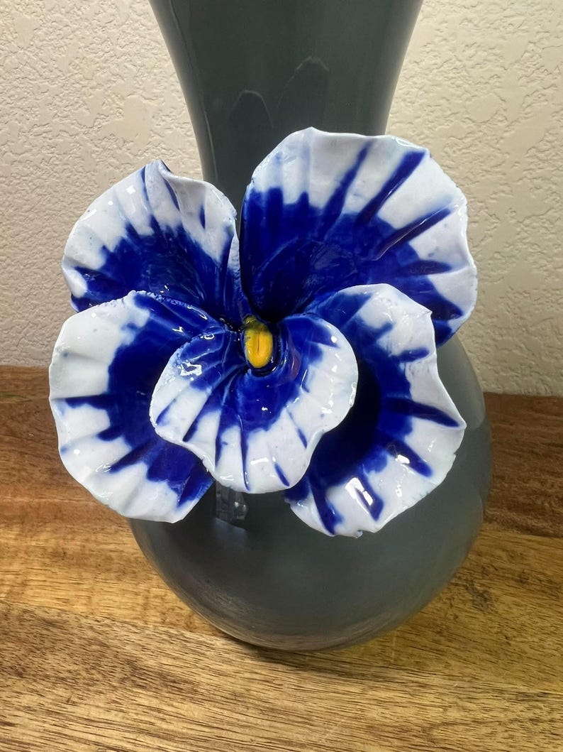 May include: A ceramic vase in a dark gray hue, adorned with a large, vibrant blue and white flower. The flower's petals display a striking contrast of colors, with a yellow center. The vase sits on a wooden surface.