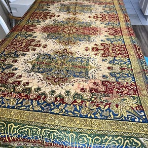 May include: A rectangular, ornate tablecloth with a cream-colored central field and a complex pattern of red, blue, green, and gold floral and geometric designs. The edges feature intricate borders and a fringed trim. The tablecloth is covering a table.