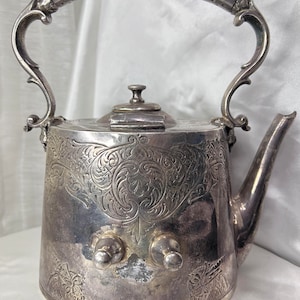 Puede incluir: Una tetera de plata con un diseño ornamentado y un asa con un adorno en espiral. La tetera tiene dos botones en la parte delantera y una tapa con un pequeño botón en la parte superior.