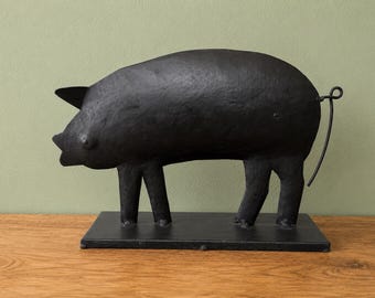 May include: A matte black metal pig sculpture standing on a rectangular base. The pig features a textured surface, pointed ears, and a thin, curled wire tail. The entire piece is finished in a solid dark color, showcasing a simple, stylized form.