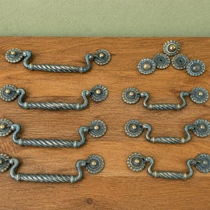May include: A collection of seven metal drawer pulls and one matching circular knob. Each pull features a twisted rope-style handle with ornate, circular mounting plates at both ends. The hardware has a dark, weathered finish with gold-toned accents.