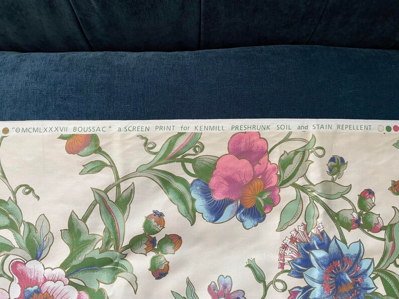 May include: Cream-colored fabric with a floral screen print. The design features large pink, blue, and purple flowers with green leaves and stems. Text at the top reads "@MCMLXXXVII BOUSSAC" and "a SCREEN PRINT for KENMILL PRESHRUNK SOIL and STAIN REPELLENT."