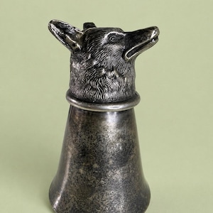 Könnte beinhalten: Ein silberfarbener Flaschenverschluss mit einem detaillierten Fuchskopf-Design. Der Fuchskopf befindet sich auf einem glockenförmigen Verschluss, der einen rustikalen Charme verleiht. Das Metall hat ein leicht gealtertes Aussehen.