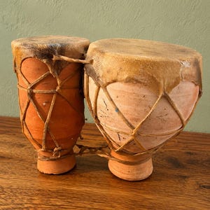 May include: A pair of connected clay drums featuring terracotta-colored bodies and natural animal hide heads. The drums are secured with a crisscross pattern of thin leather lacing that wraps around the exterior of both vessels to hold the heads in place.