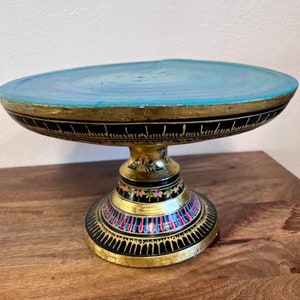 May include: A decorative cake stand with a round, turquoise top and a gold rim. The black base is adorned with gold and colorful floral patterns. The stand is made of wood and is suitable for displaying desserts or other items.