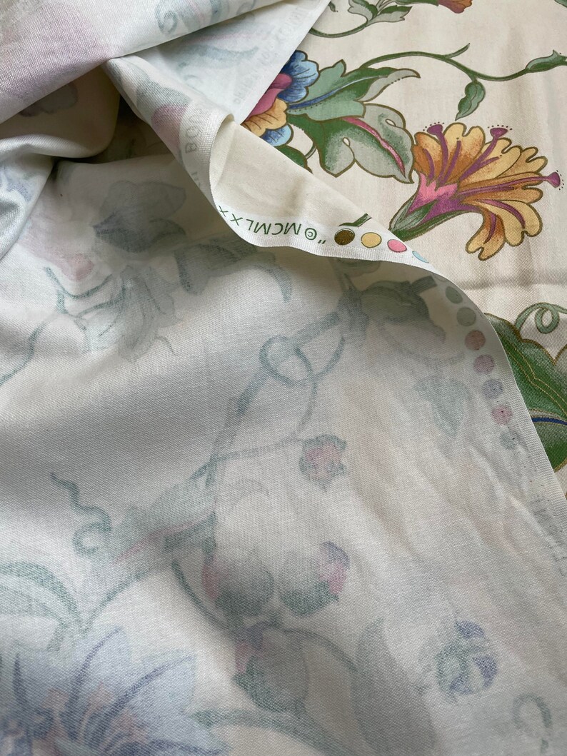 May include: Cream-colored fabric with a floral pattern featuring large, stylized flowers in shades of yellow, pink, blue, and green. The fabric is folded, revealing the underside with a softer, more muted floral design. The text "&copy; MCMLXX" is visible.