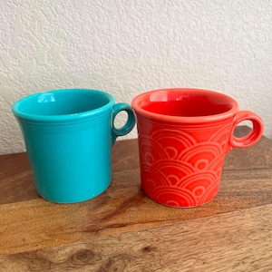May include: A solid turquoise ceramic mug with a smooth finish and a matching handle sits next to a bright orange ceramic mug featuring a raised, repeating semi-circular wave pattern and a circular handle. Both mugs have a flared rim design.