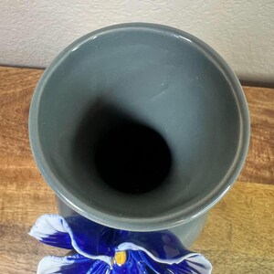 May include: A gray ceramic vase with a flared top and a large, detailed blue and white pansy flower accenting the base. The flower has a yellow center. The vase is set against a wooden surface.