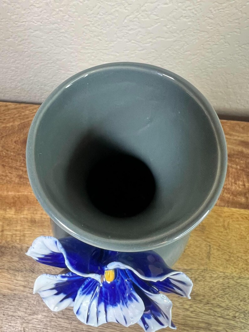 May include: A gray ceramic vase with a flared top and a large, detailed blue and white pansy flower accenting the base. The flower has a yellow center. The vase is set against a wooden surface.