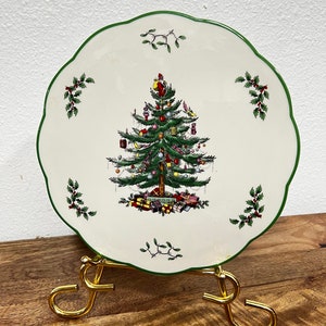 May include: A white ceramic plate with a green rim features a Christmas tree design with red, green, and gold ornaments. The plate is on a gold metal stand.