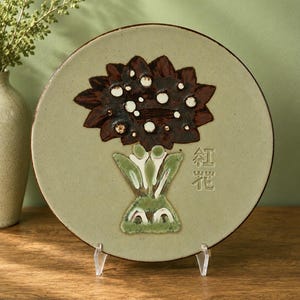 Pode incluir: Um prato decorativo de cerâmica com um design floral. O prato apresenta uma flor marrom com detalhes brancos e um caule verde. O prato tem caracteres japoneses e é exibido em um suporte transparente.