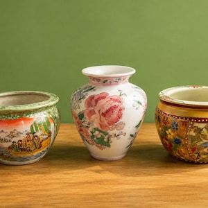 May include: Three decorative ceramic vases. The center vase is white with a pink floral design. The vase on the left is green with a landscape scene. The vase on the right is yellow with a floral and bird design.
