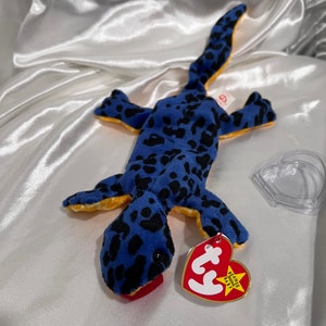 May include: A blue plush lizard toy with black spots and yellow accents. The toy has a red mouth and a red heart-shaped tag that reads "Beanie Baby". The lizard is lying on a white satin surface.