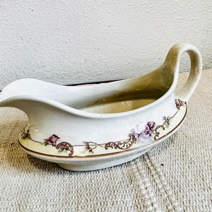 Pode incluir: Uma molheira de cerâmica branca com um design floral em tons de rosa, verde e marrom. A molheira tem uma alça e uma base larga e rasa.