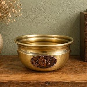 May include: A polished brass bowl featuring a rounded shape and a wide, flared rim. Centered on the front of the bowl is an oval-shaped copper-toned inlay displaying a relief design of a single flower with leaves and stems.