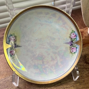 May include: A small, round, antique porcelain plate with a gold rim and iridescent finish. The plate features hand-painted pink and purple roses on each side. The plate is displayed on a clear acrylic stand.
