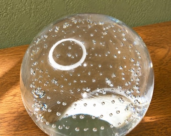May include: A clear glass sphere paperweight featuring a multitude of small, suspended air bubbles throughout its interior. The spherical object has a smooth, polished surface that reflects light, creating a bright circular highlight on its top.