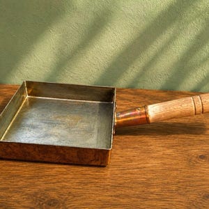 May include: A square copper pan with a metallic interior and a long, cylindrical wooden handle attached by a copper ferrule. The exterior of the pan is polished copper, while the interior shows a silver-toned finish with visible signs of use.