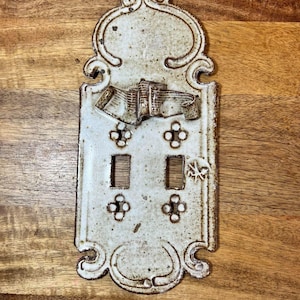May include: A rectangular ceramic light switch plate with a speckled off-white glaze and brown edges. The plate features a decorative molded ribbon at the top, two rectangular cutouts for switches, and four small circular screw hole openings.