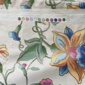 May include: Cream-colored fabric with a floral pattern featuring large flowers in shades of orange, blue, and pink, with green leaves and stems. A color swatch with various pastel colors is visible. The fabric has a subtle sheen.