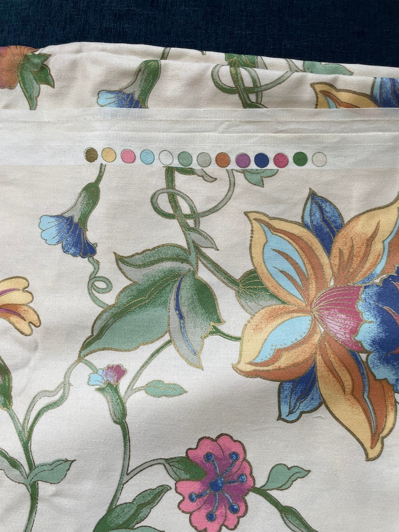 May include: Cream-colored fabric with a floral pattern featuring large flowers in shades of orange, blue, and pink, with green leaves and stems. A color swatch with various pastel colors is visible. The fabric has a subtle sheen.