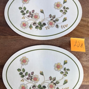 Può includere: Due tovagliette ovali bianche con un bordo verde. Le tovagliette presentano un motivo floreale con fiori bianchi e gialli e foglie verdi.